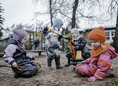 Fire børn sidder i flyverdragt i en sandkasse og leger.