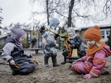 Fire børn sidder i flyverdragt og leger i en sandkasse